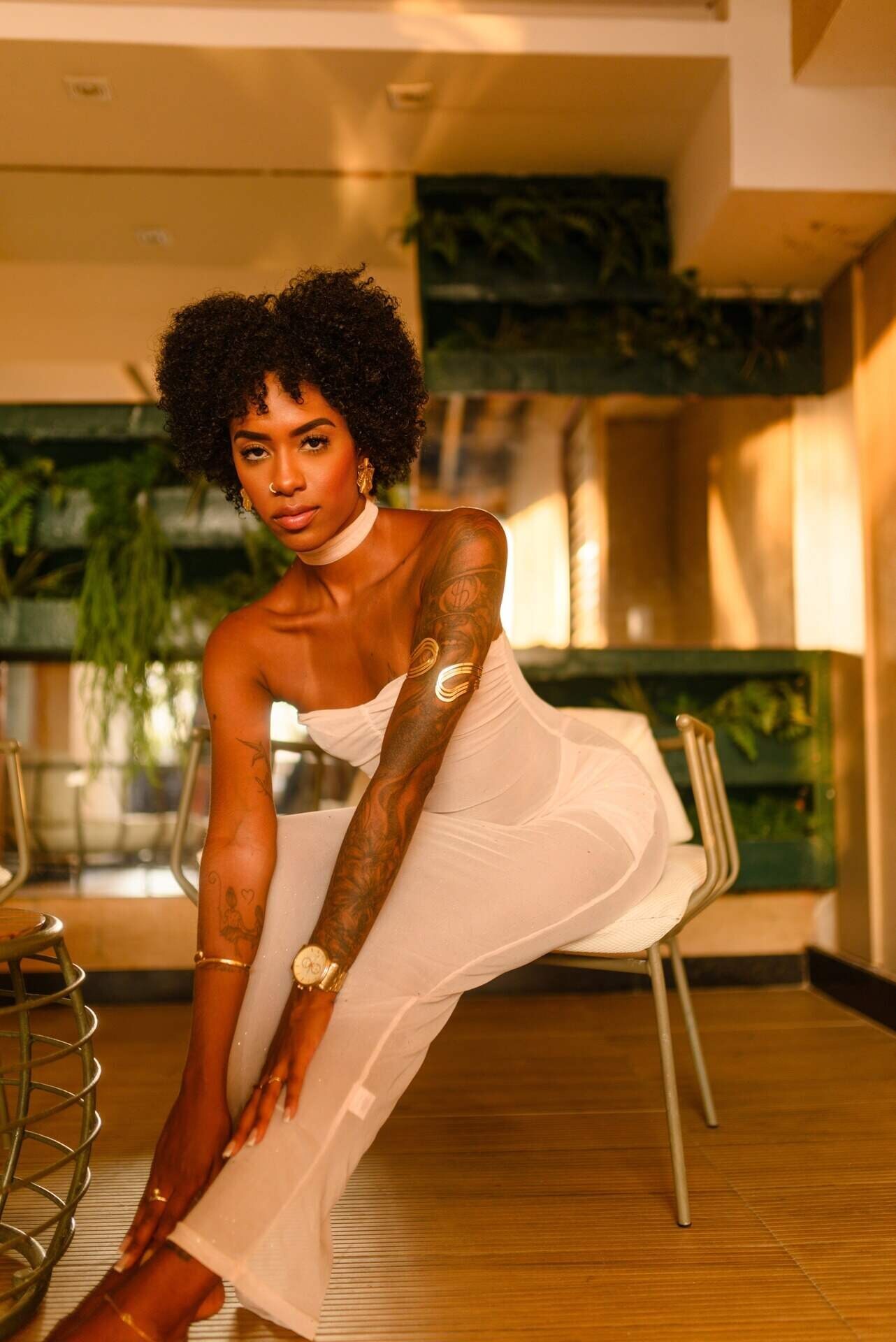 Tattooed Black woman with afro in white outfit sitting indoors.