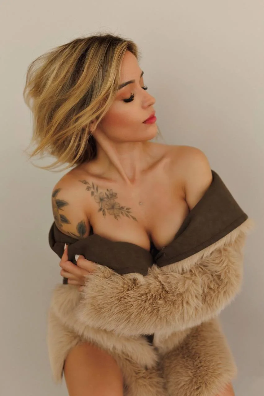 Blonde woman with floral tattoos wearing a faux fur coat.