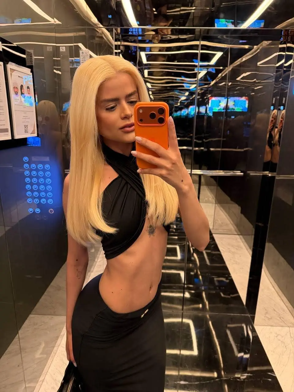 Blonde woman in black outfit taking a lift selfie.