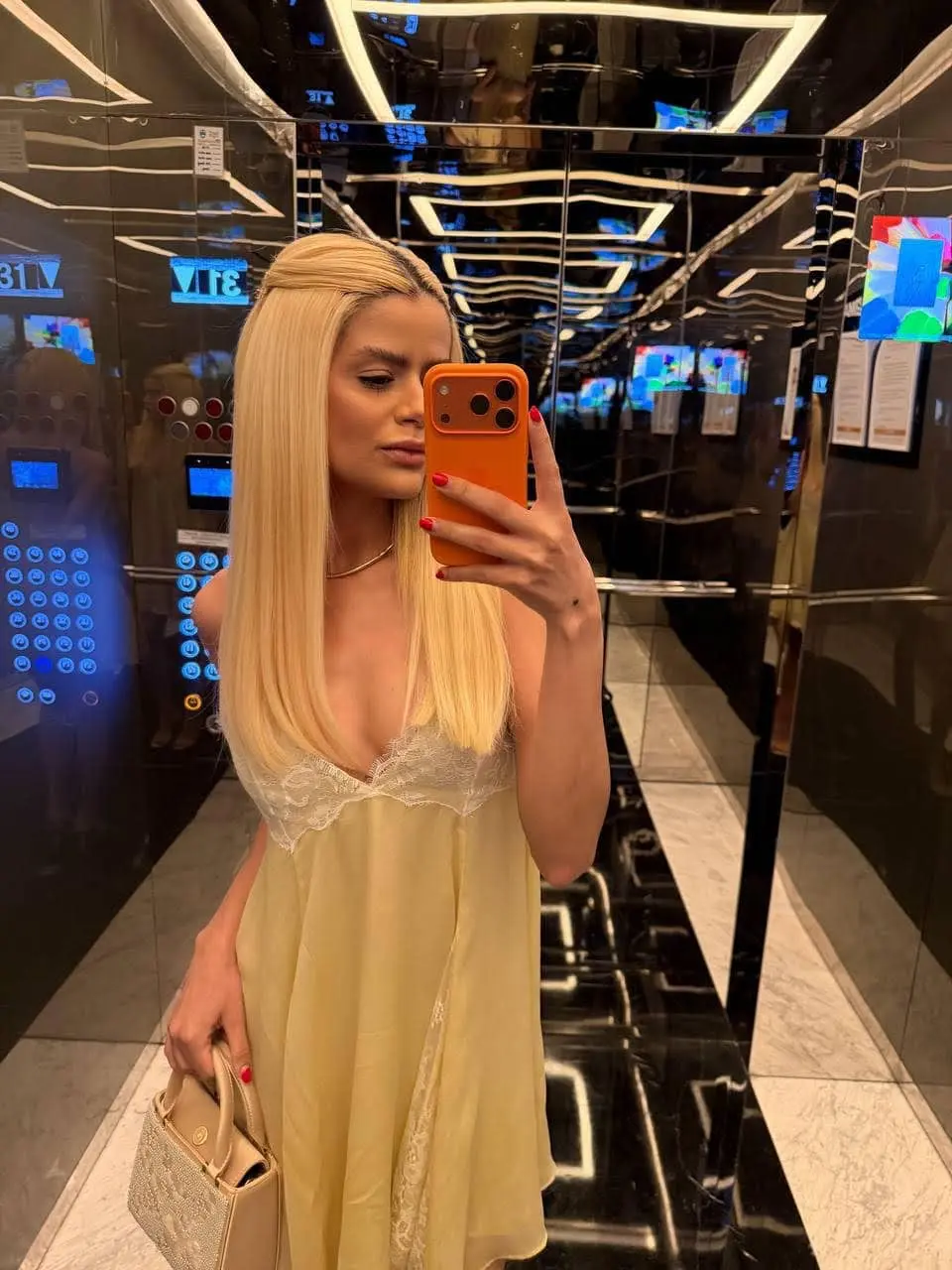 Blonde woman in a yellow slip dress taking lift selfie.