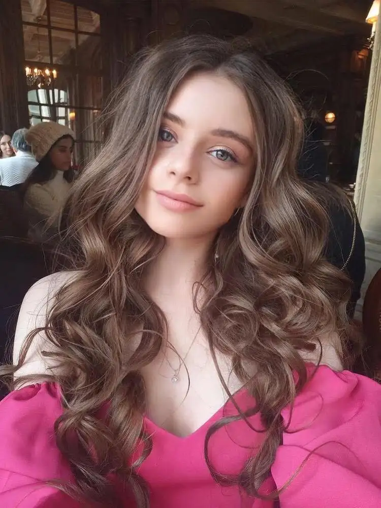 Young woman with long, wavy brown hair wearing pink top.