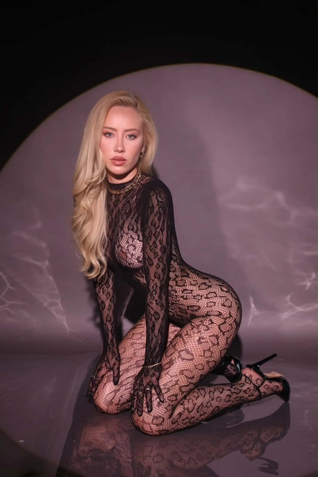 Blonde woman in patterned mesh bodysuit kneeling in studio.