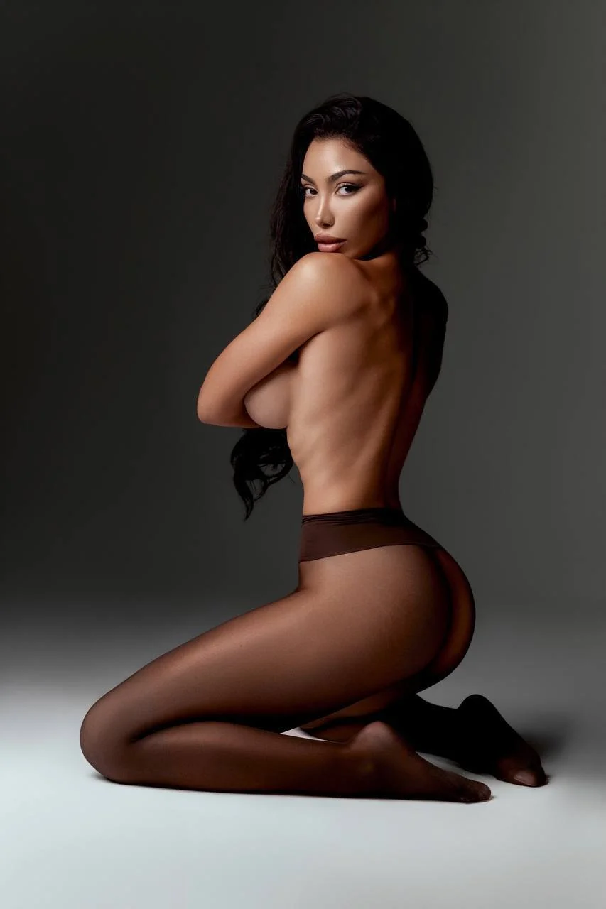 Topless woman kneeling in dark tights, looking over her shoulder.