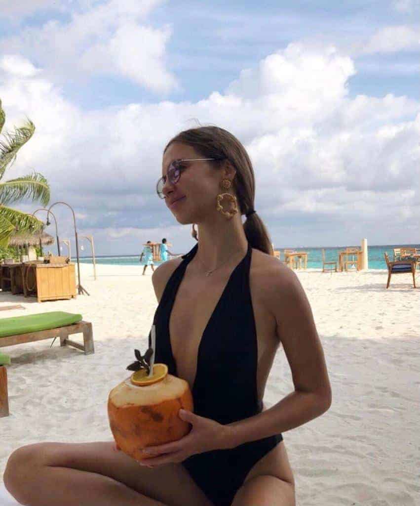 Woman in black swimsuit holding coconut on beach.
