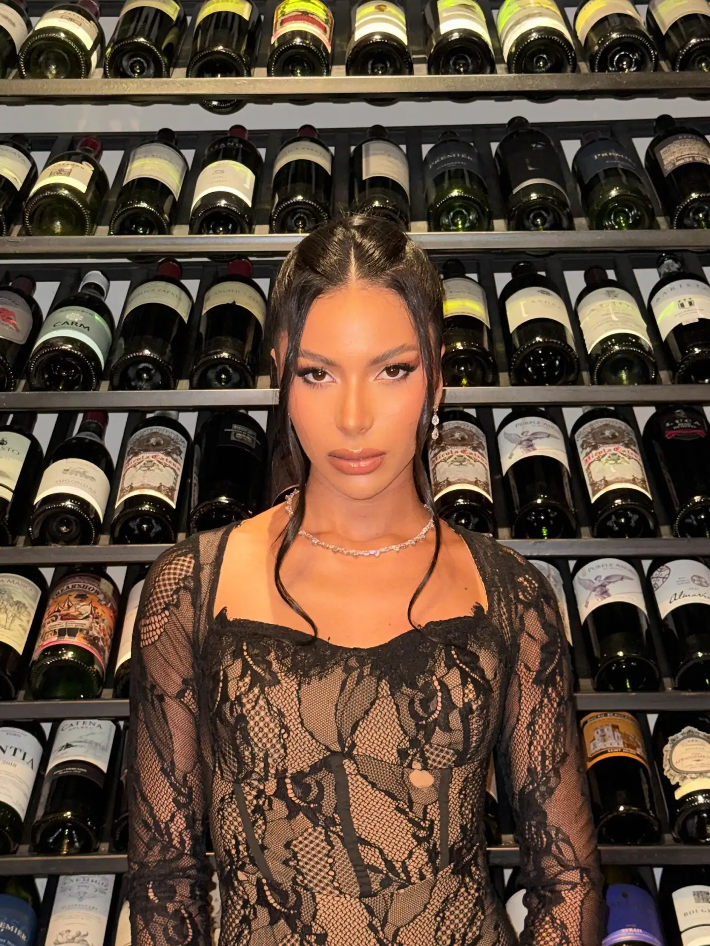Woman in black lace top posing before a wine rack.