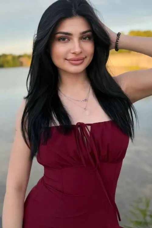 Woman in maroon dress posing by a tranquil lake.