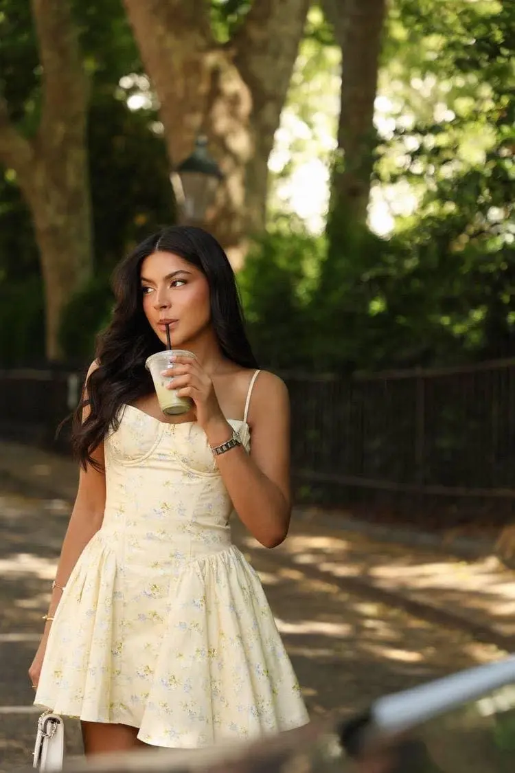 Woman in yellow floral dress sipping an iced beverage outdoors.