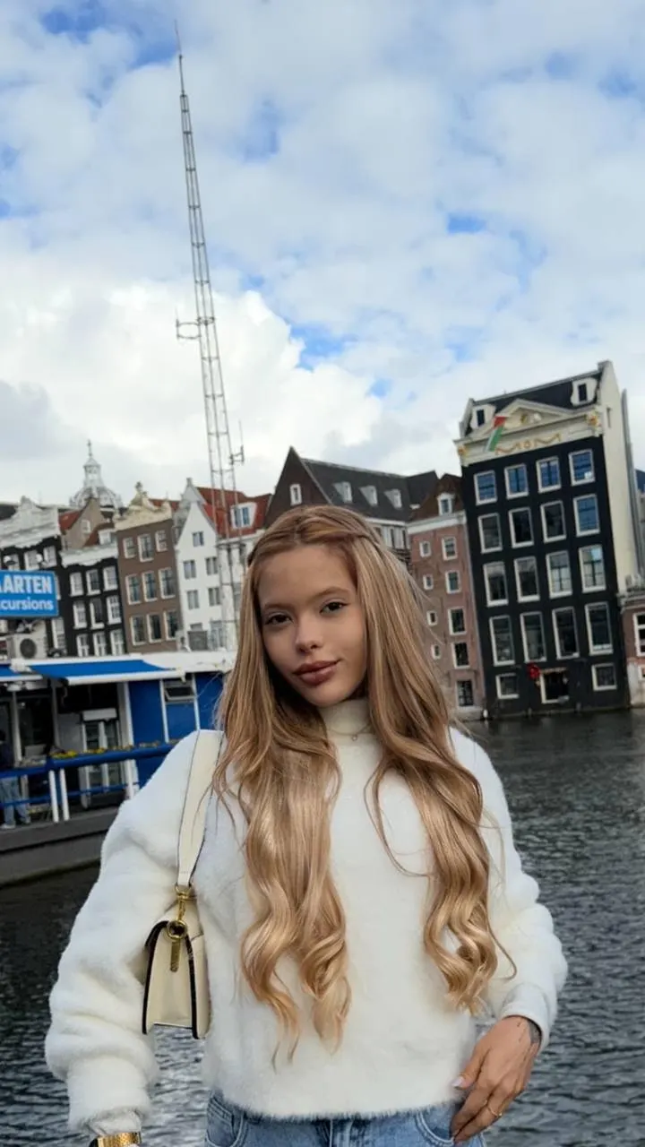 Blonde woman in white jumper posing before Amsterdam canal houses.