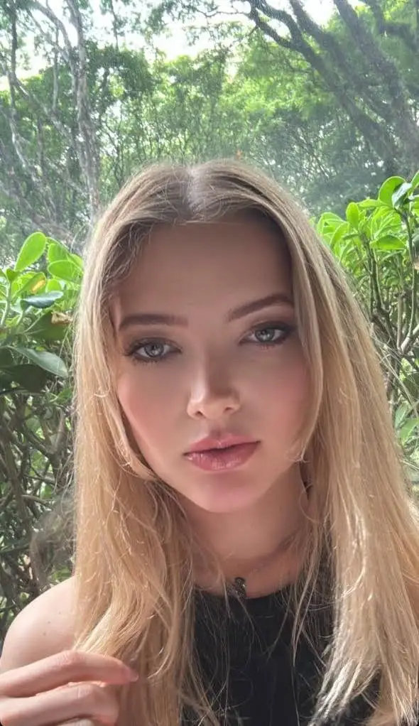 A young woman with blonde hair outdoors against green trees.
