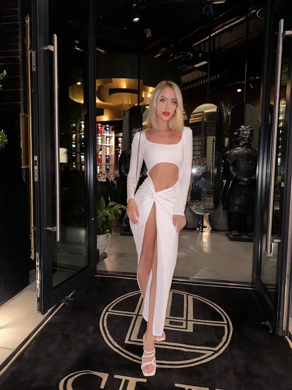 Blonde woman in white cut-out dress at restaurant entrance.