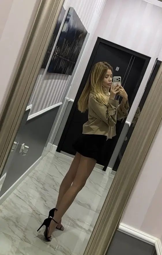 Woman posing in a tan jacket, black skirt, and heels.