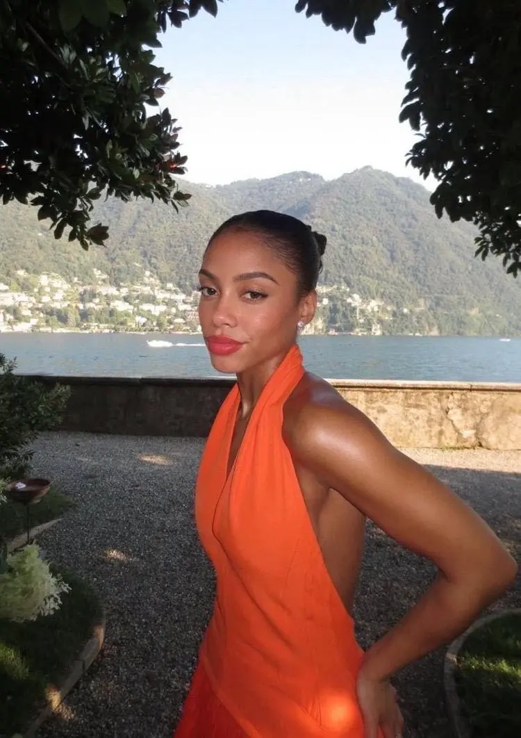 Woman in an orange halter dress posing before a lake.
