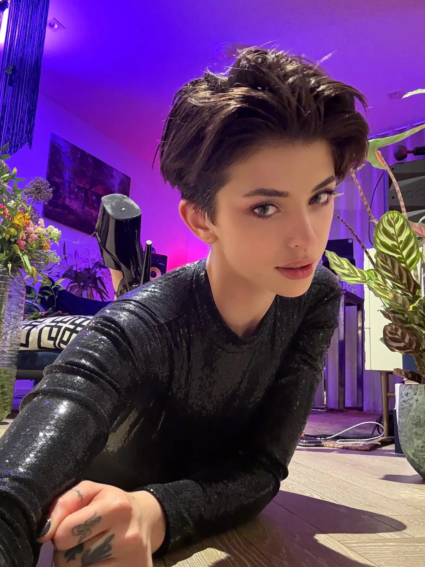 Person with pixie cut in black sequinned top, purple lighting.
