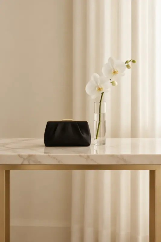 A black satin evening clutch with a gold clasp and a single white orchid in a glass vase on a marble console, in front of cream curtains.