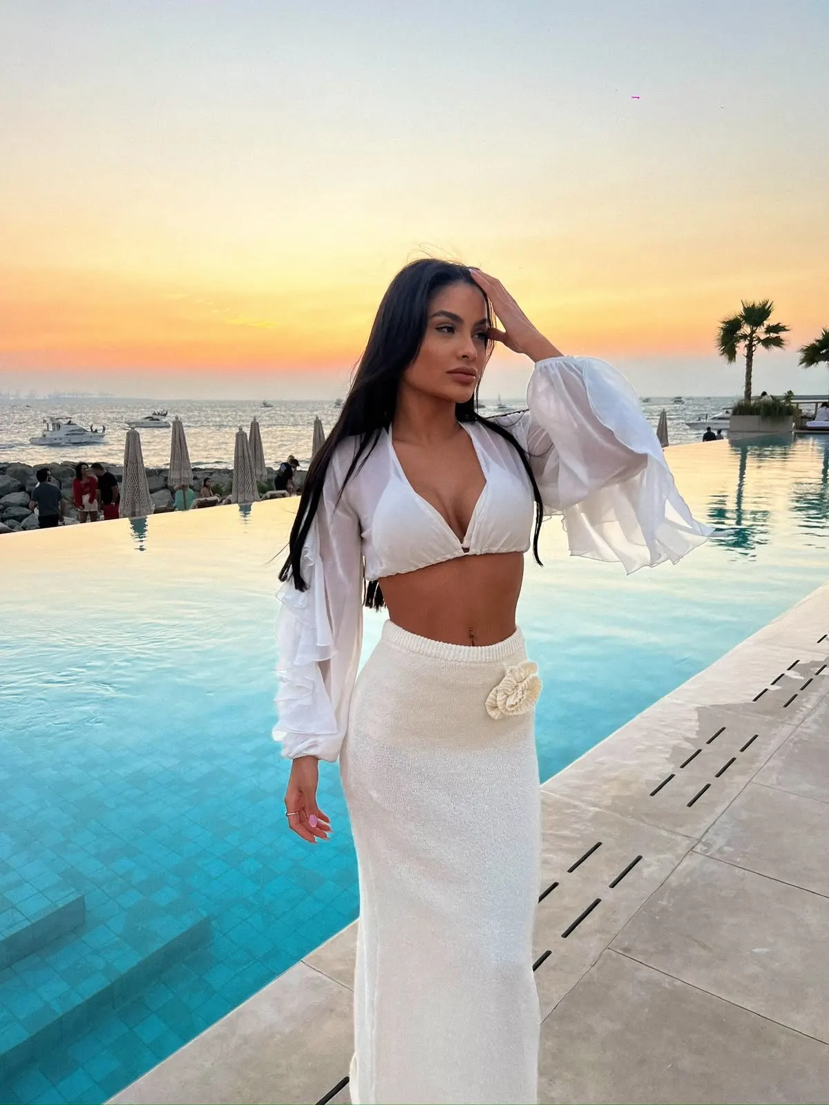 Woman in white outfit posing by infinity pool at sunset.