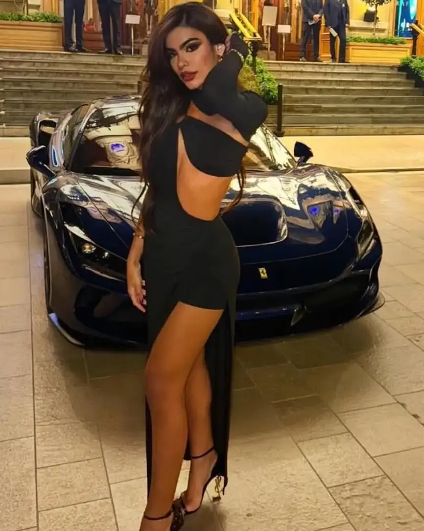 Woman in black dress posing beside a blue Ferrari supercar.