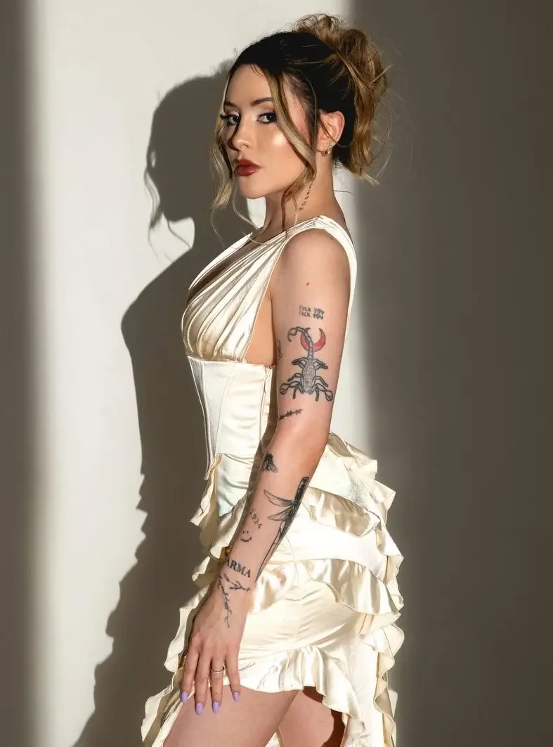 Tattooed woman in cream ruffled dress posing against a wall.