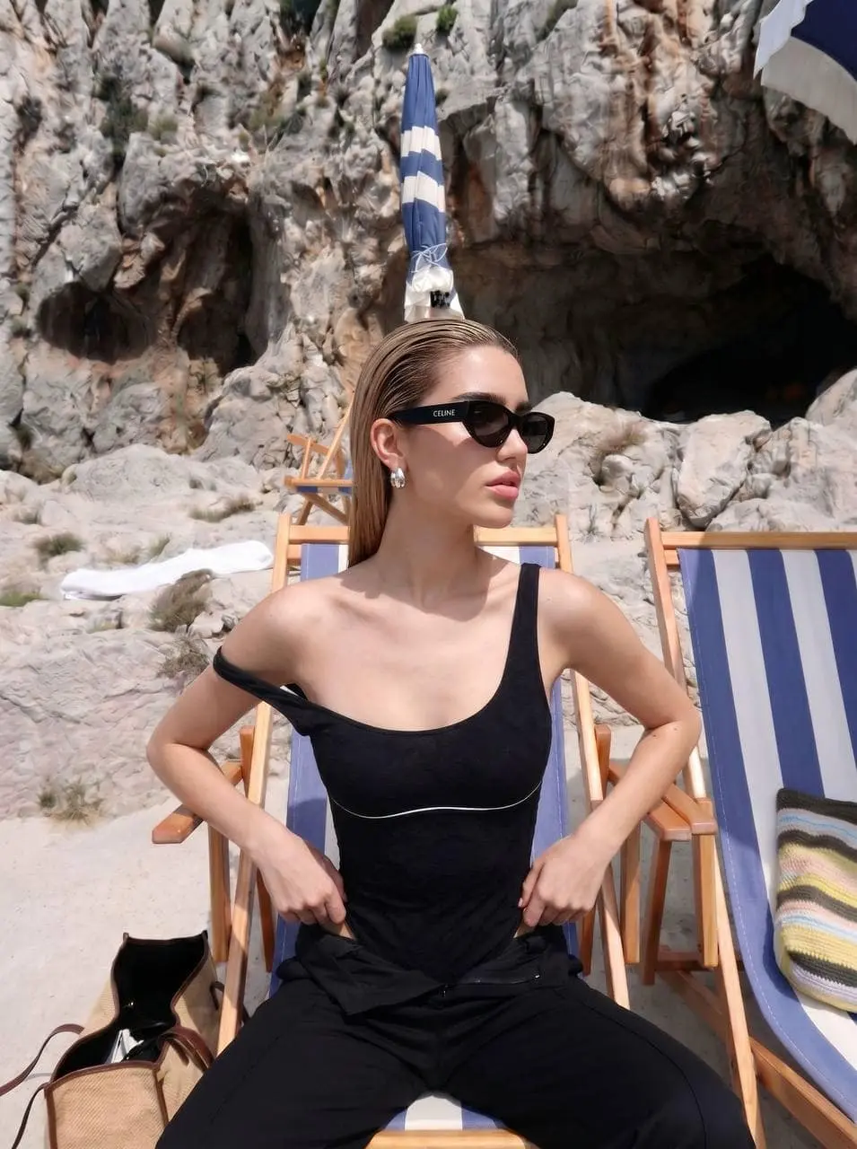 Woman in black swimsuit and sunglasses at a rocky beach.