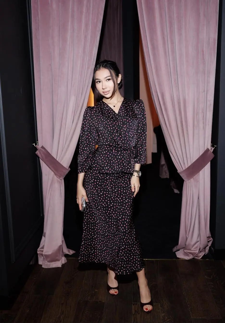 Woman in floral dress standing between pink velvet curtains.