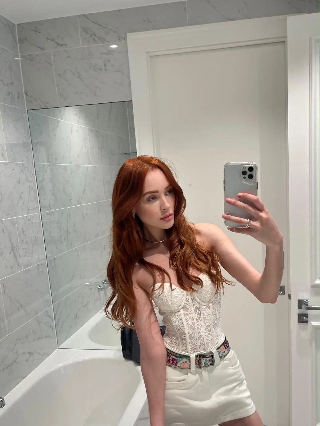 Red-haired woman in white lace corset taking a mirror selfie.