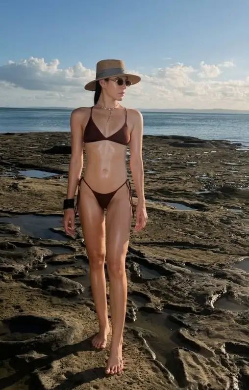 Woman in brown bikini, straw hat, and sunglasses on rocks.