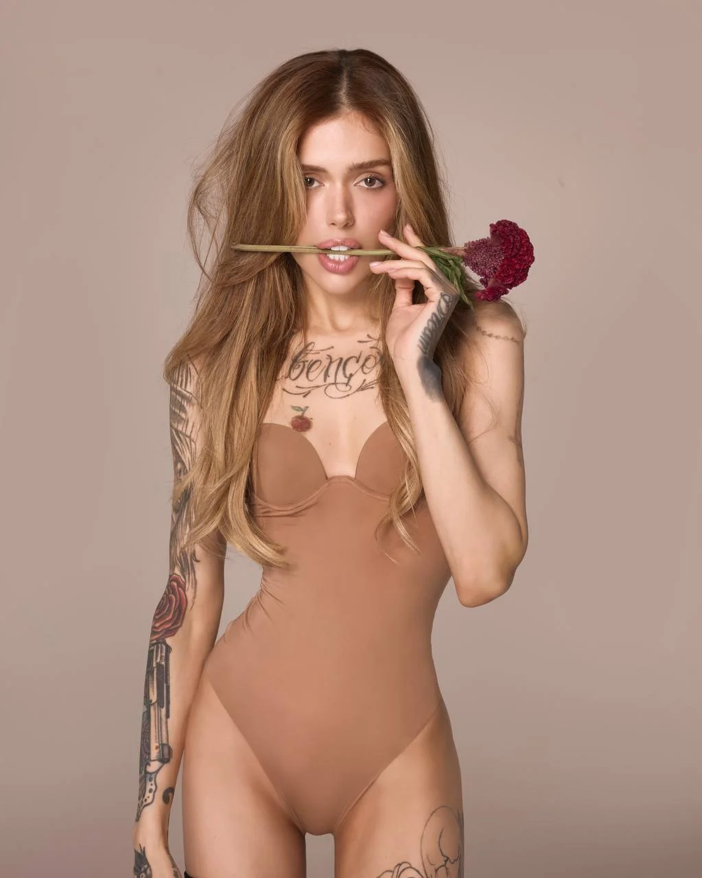 Tattooed woman in a beige bodysuit holding a red flower.